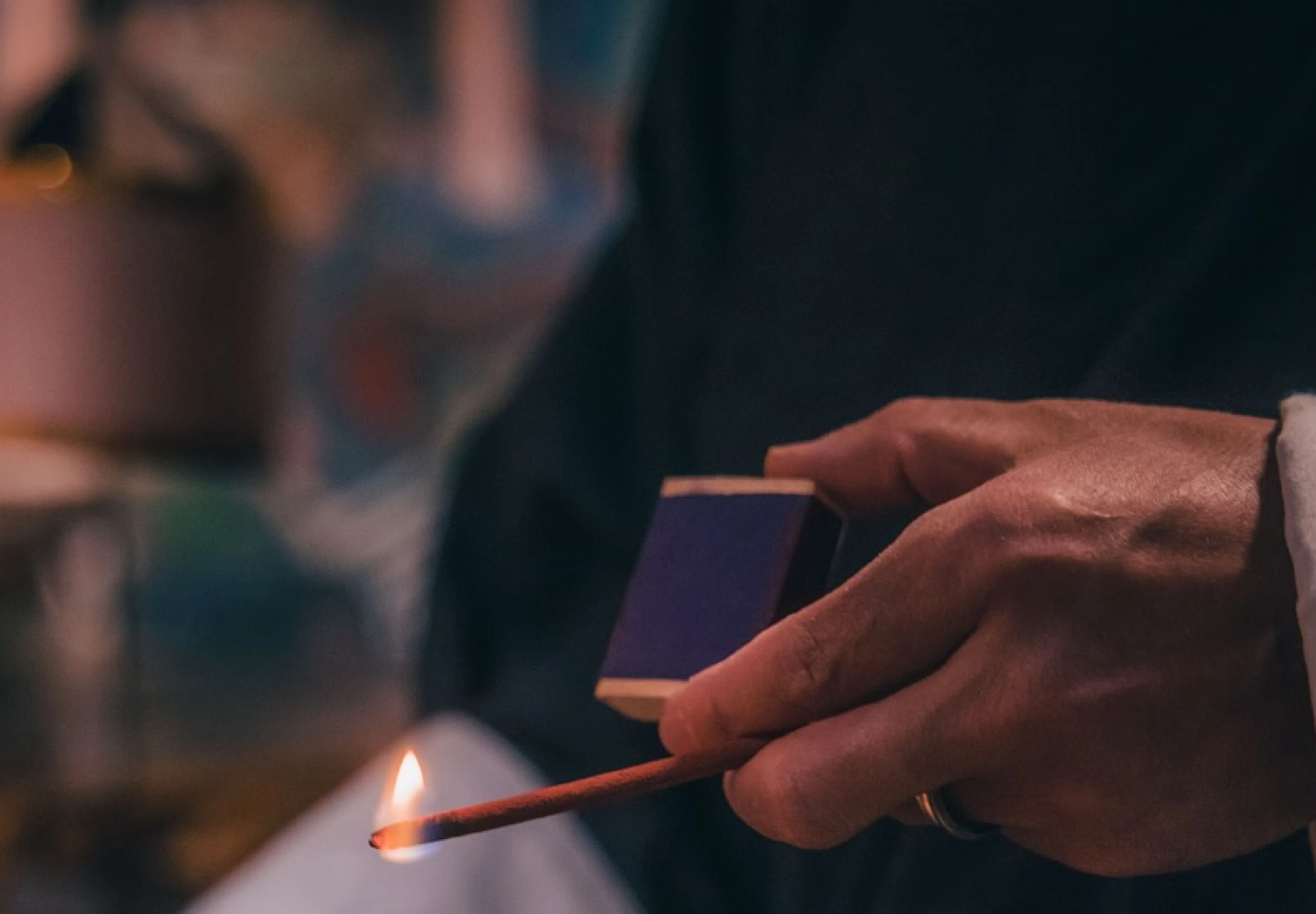 Hand lighting a match beside a candle in a dim, atmospheric room
